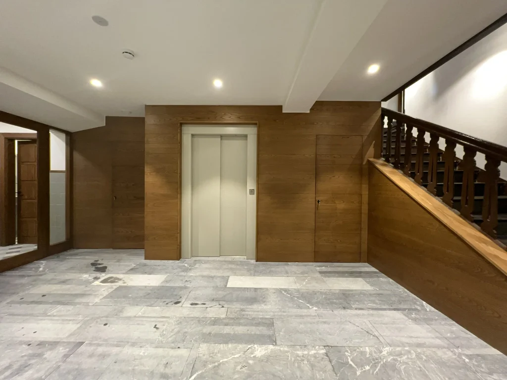 Vestíbulo con solado de piedra, ascensor forrado de madera y escalera